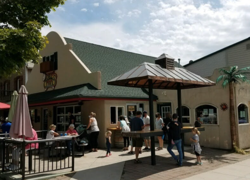 A busy outdoor area of a restaurant with people dining, a shaded seating area, and a palm tree decoration.