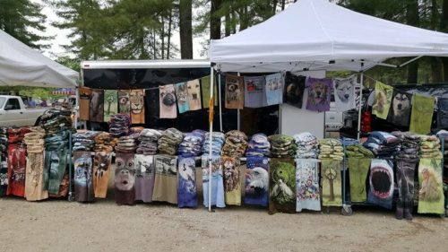 A market stall displaying a variety of colorful blankets featuring animal designs, with tents and trees in the background.