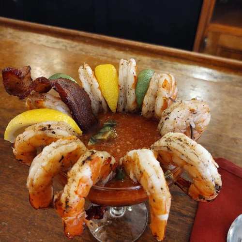A cocktail glass filled with shrimp, garnished with lemon wedges and bacon, served on a wooden bar.
