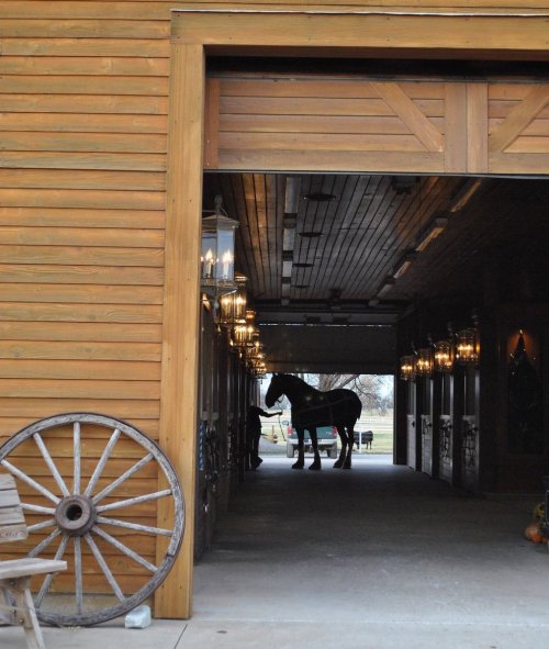 A wooden barn entrance with a horse visible inside, flanked by lanterns and a wagon wheel in the foreground.