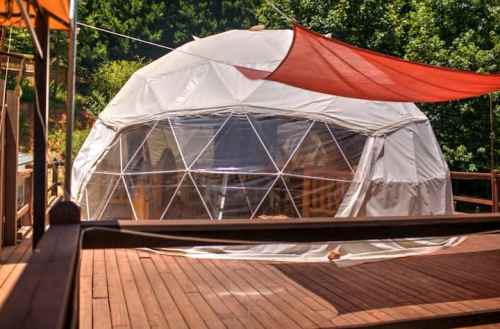 A geodesic dome on a wooden deck, surrounded by greenery and shaded by colorful fabric canopies.