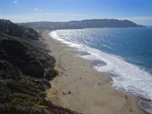 A scenic beach view with gentle waves, sandy shore, and people enjoying the sun under a clear blue sky.