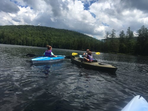 Two people kayaking on a calm lake surrounded by green trees and a cloudy sky.