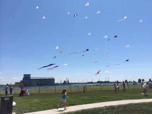 Children fly colorful kites on a sunny day, with a clear blue sky and a distant industrial backdrop.