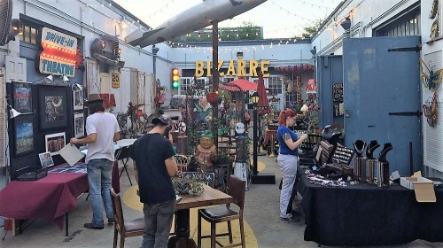 A vibrant market scene with vendors displaying crafts and art in a lively, decorated outdoor space.