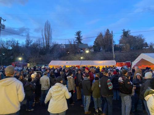 A large crowd gathers at an outdoor event in the evening, with tents and trees in the background under a blue sky.