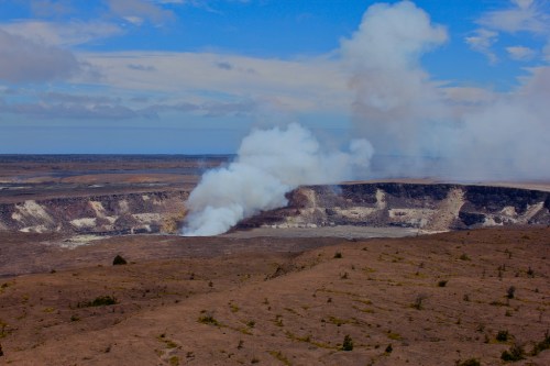 A volcanic crater with smoke rising, surrounded by a barren landscape under a blue sky with scattered clouds.