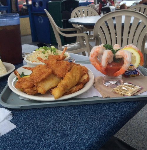 A tray with fried fish, shrimp cocktail, and coleslaw, set on a blue table in a casual dining area.