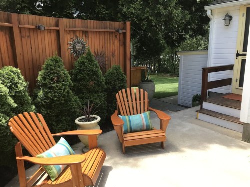 Two wooden chairs with cushions sit on a patio surrounded by greenery and a wooden fence with a sun decoration.