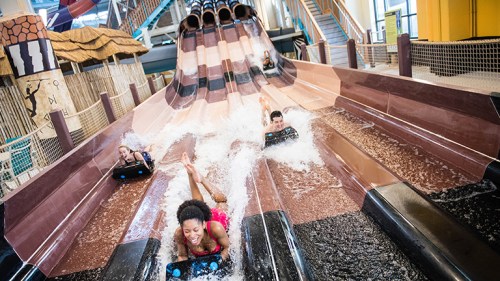 Four people slide down a water slide, splashing into the pool below in a vibrant indoor water park.