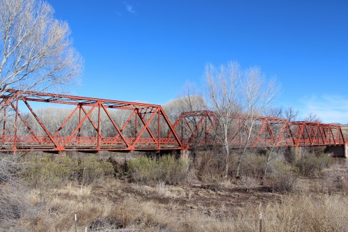 A red metal truss bridge spans a dry riverbed, surrounded by sparse trees and blue sky.