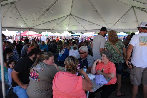 A crowded outdoor event under a large tent, with people socializing at tables and colorful tents in the background.