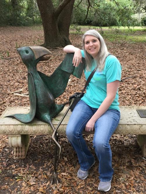 A woman in a teal shirt sits beside a bronze bird sculpture on a stone bench in a wooded area.
