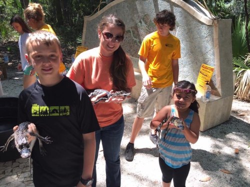 Two children hold snakes while a woman assists them; a teen stands nearby in a nature setting with warning signs.