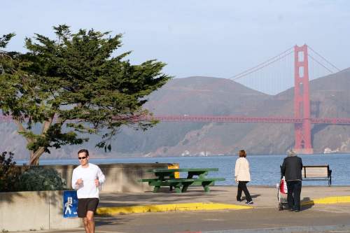 A jogger runs along a waterfront path with the Golden Gate Bridge in the background and people walking nearby.