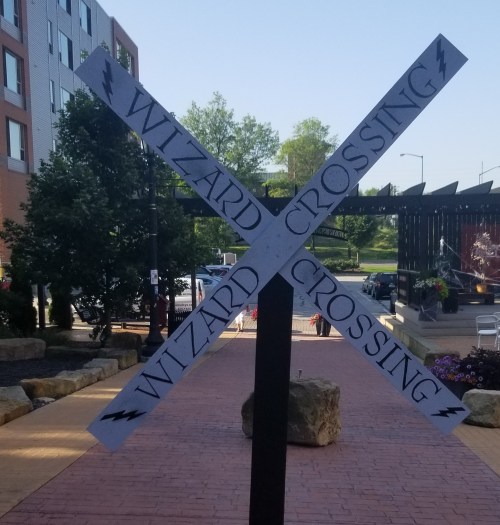 Signpost with "Wizard Crossing" written in four directions, set in a landscaped area with buildings and flowers nearby.