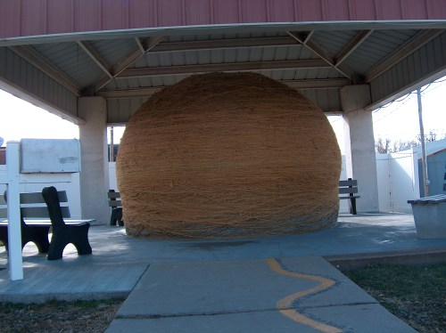 A large, spherical ball of twine sits under a shelter, with benches and a pathway visible in the foreground.