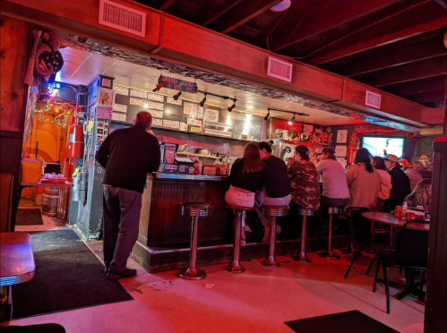 A dimly lit bar scene with patrons sitting at the counter, colorful lights, and a cozy atmosphere.