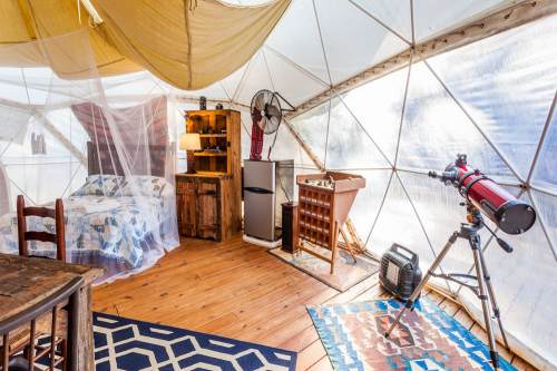Cozy glamping tent interior with a bed, furniture, telescope, and decorative rugs. Natural light filters through.