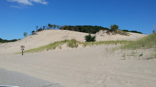A sandy dune with sparse vegetation under a clear blue sky, alongside a road with a sign visible in the foreground.