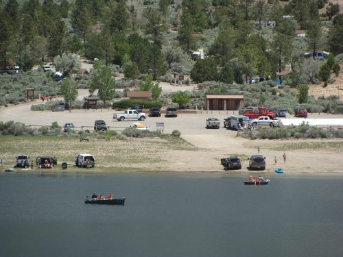 A scenic view of a lakeside area with parked vehicles, campers, and people boating on the water.