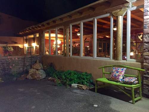 A cozy outdoor seating area at night, featuring a green bench with a colorful pillow and a well-lit windowed room.