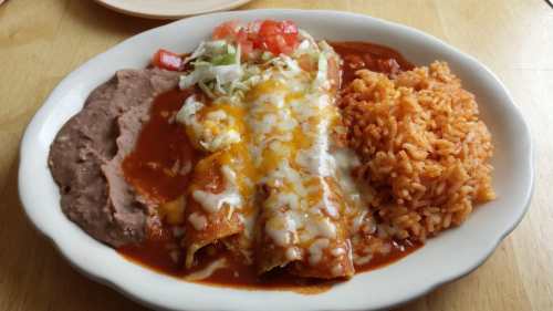 A plate of enchiladas topped with cheese and sauce, served with refried beans, rice, and fresh toppings.