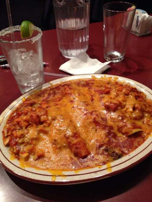A plate of cheesy enchiladas topped with melted cheese, served with a glass of water and a cocktail.