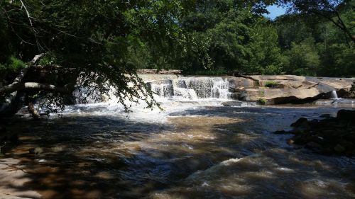 A serene river scene featuring a waterfall cascading over rocks, surrounded by lush greenery and a clear blue sky.