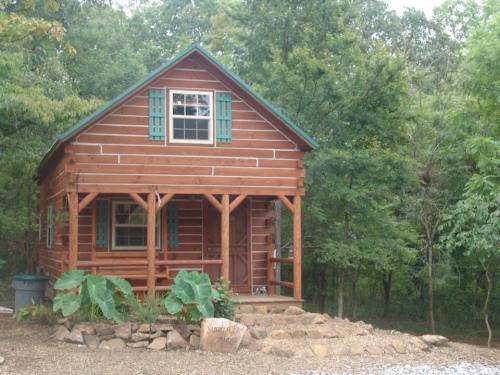 A small wooden cabin with green shutters, surrounded by trees and a gravel path.