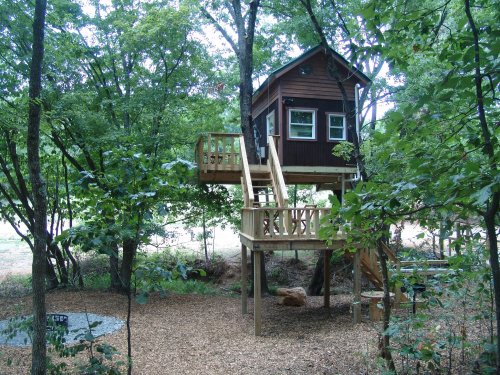 A wooden treehouse elevated among green trees, featuring a deck and stairs, surrounded by a natural landscape.