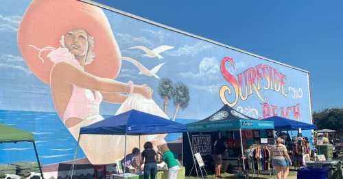 A colorful mural of a woman in a large hat at Surfside Beach, with market tents and people in the foreground.