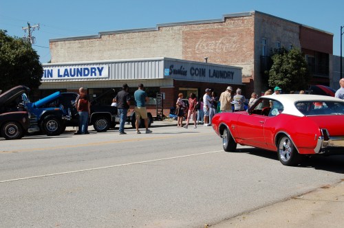 A red classic car parked on the street near a busy coin laundry with people gathered outside.