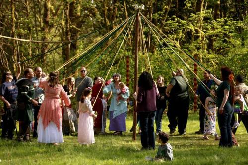 A group of people in colorful costumes gather around a maypole in a forest clearing, celebrating together.