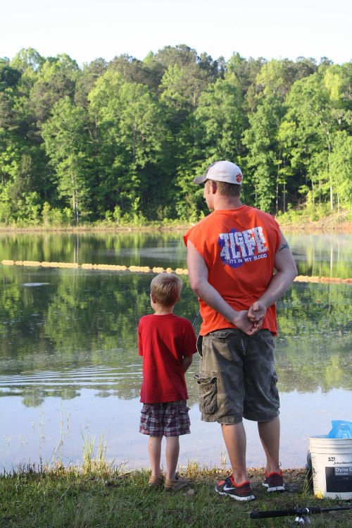 A man and a boy stand by a calm lake, watching the water surrounded by lush green trees.