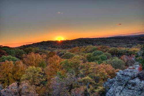 A vibrant sunset over a forested landscape, showcasing autumn colors and a clear sky.