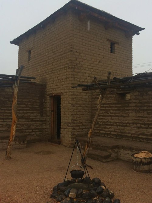 A traditional adobe structure with a tower, surrounded by a stone circle and wooden supports, under a cloudy sky.