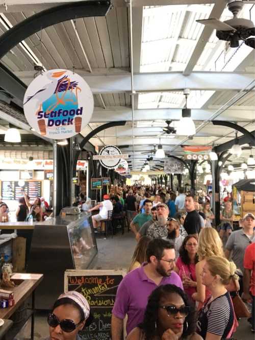 A bustling market scene with people browsing food stalls under a high ceiling, featuring signs for seafood and other vendors.