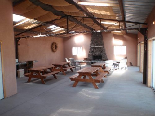 Interior of a spacious room with wooden picnic tables, a stone fireplace, and large windows, featuring a rustic design.