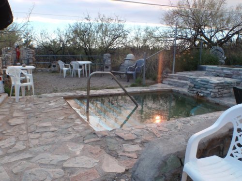 A small, tranquil hot spring surrounded by stone pathways and white chairs, with trees in the background.