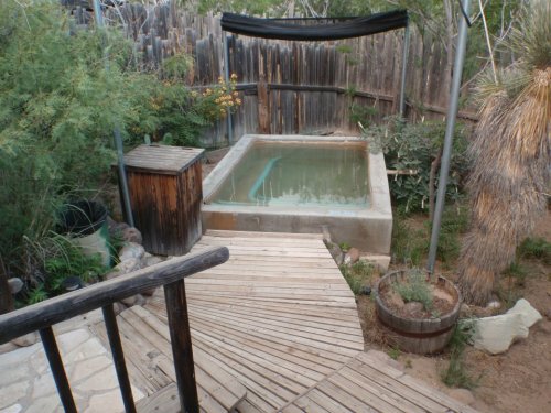 A wooden deck leads to a small, empty hot tub surrounded by greenery and rustic fencing.