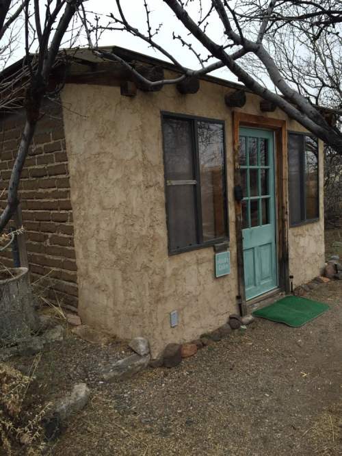 A small, rustic adobe-style cabin with a turquoise door, surrounded by bare trees and gravel.