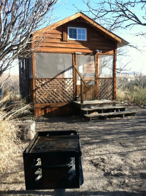A small wooden cabin with a screened porch, surrounded by dry grass and trees, with a grill in the foreground.
