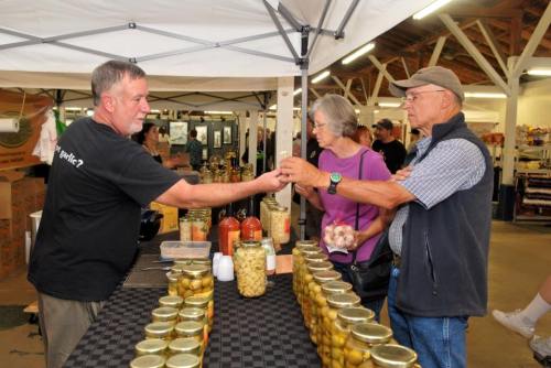 A vendor at a market hands a jar to two customers, surrounded by jars of pickles and other goods on the table.