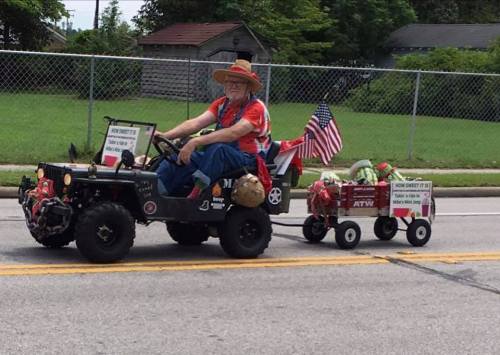 An elderly man in a straw hat drives a decorated mini vehicle, pulling a wagon with signs and festive decorations.