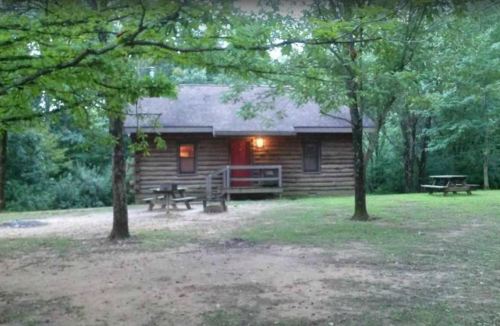 A rustic log cabin surrounded by trees, featuring a red door and picnic tables in a grassy area.