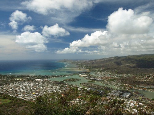 A panoramic view of a coastal landscape with blue waters, green hills, and scattered clouds in the sky.
