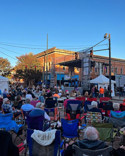 A crowd of people sitting in folding chairs at an outdoor concert, with a stage and buildings in the background.