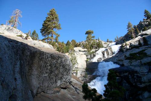 A scenic view of a waterfall cascading over rocks, surrounded by trees and a clear blue sky.