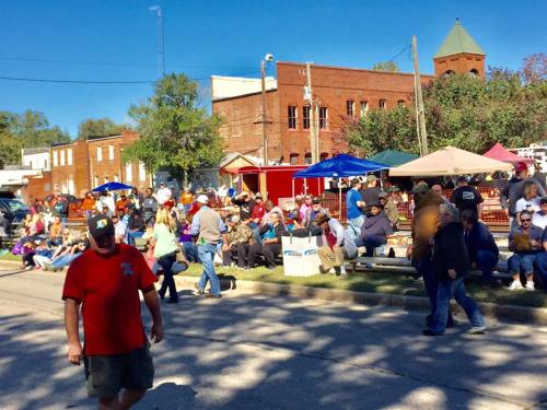 A lively street scene with a crowd enjoying a festival, featuring tents, people sitting, and a clear blue sky.